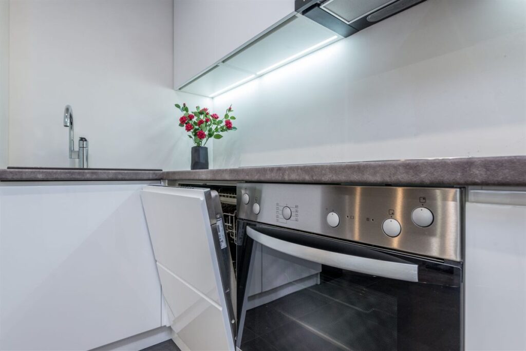 Built-in dishwasher with its door slightly open next to a wall oven in a white kitchen